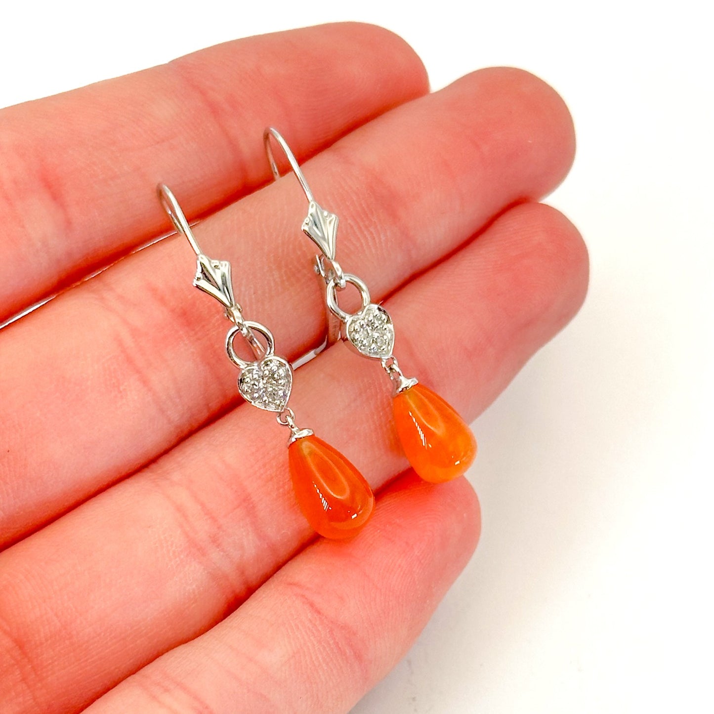 A pair of 14K white gold dangle lever-back earrings on a hand, featuring teardrop-shaped fire opals with heart-shaped and diamond accents accents on a white background.