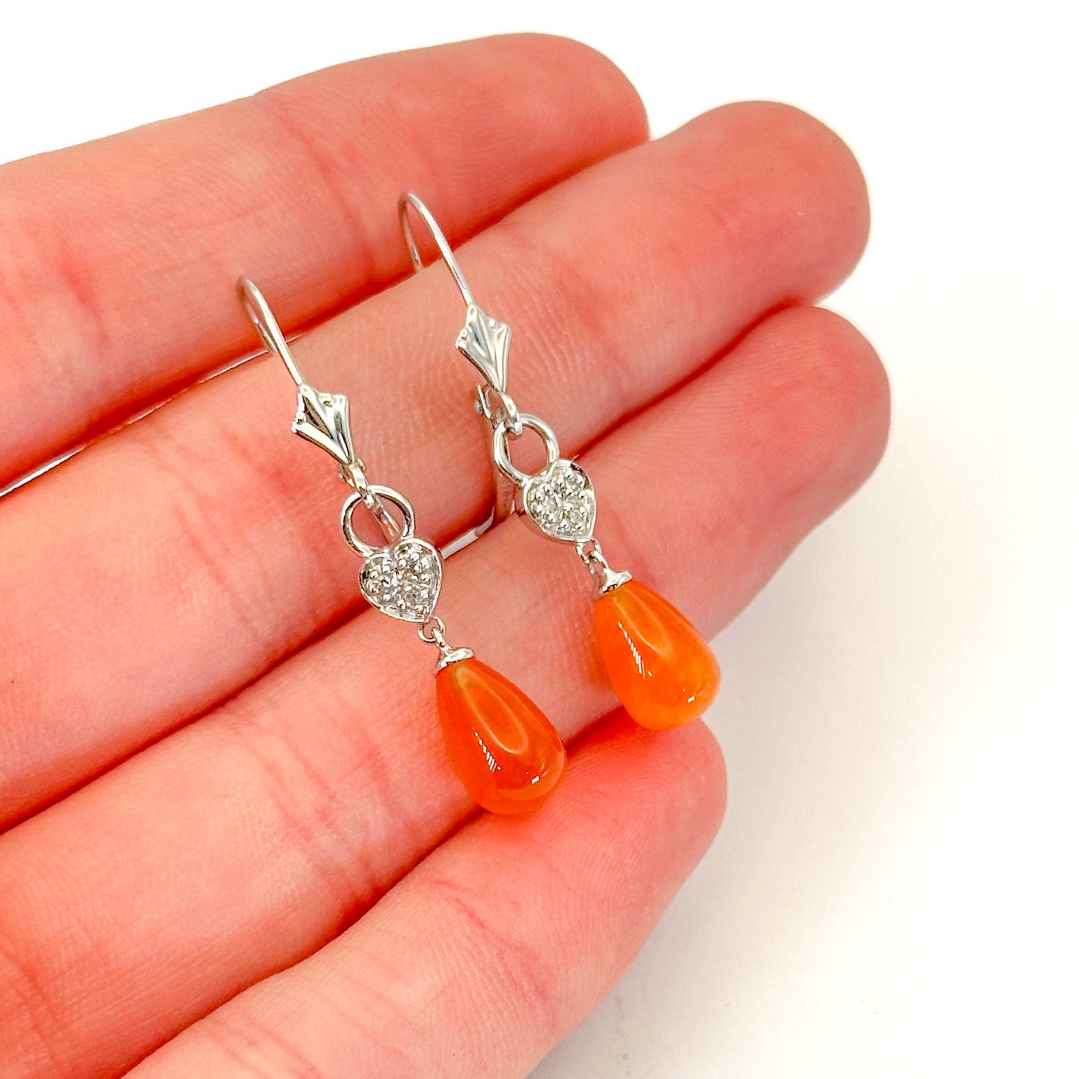 A pair of 14K white gold dangle lever-back earrings on a hand, featuring teardrop-shaped fire opals with heart-shaped and diamond accents accents on a white background.